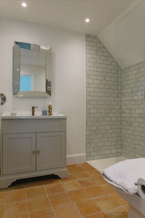 Casa de banho com toucador branco, espelho, chuveiro com porta de vidro e chão em azulejos.