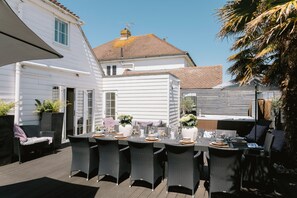 A patio with a table set for six, surrounded by wicker chairs, and a white building in the background.