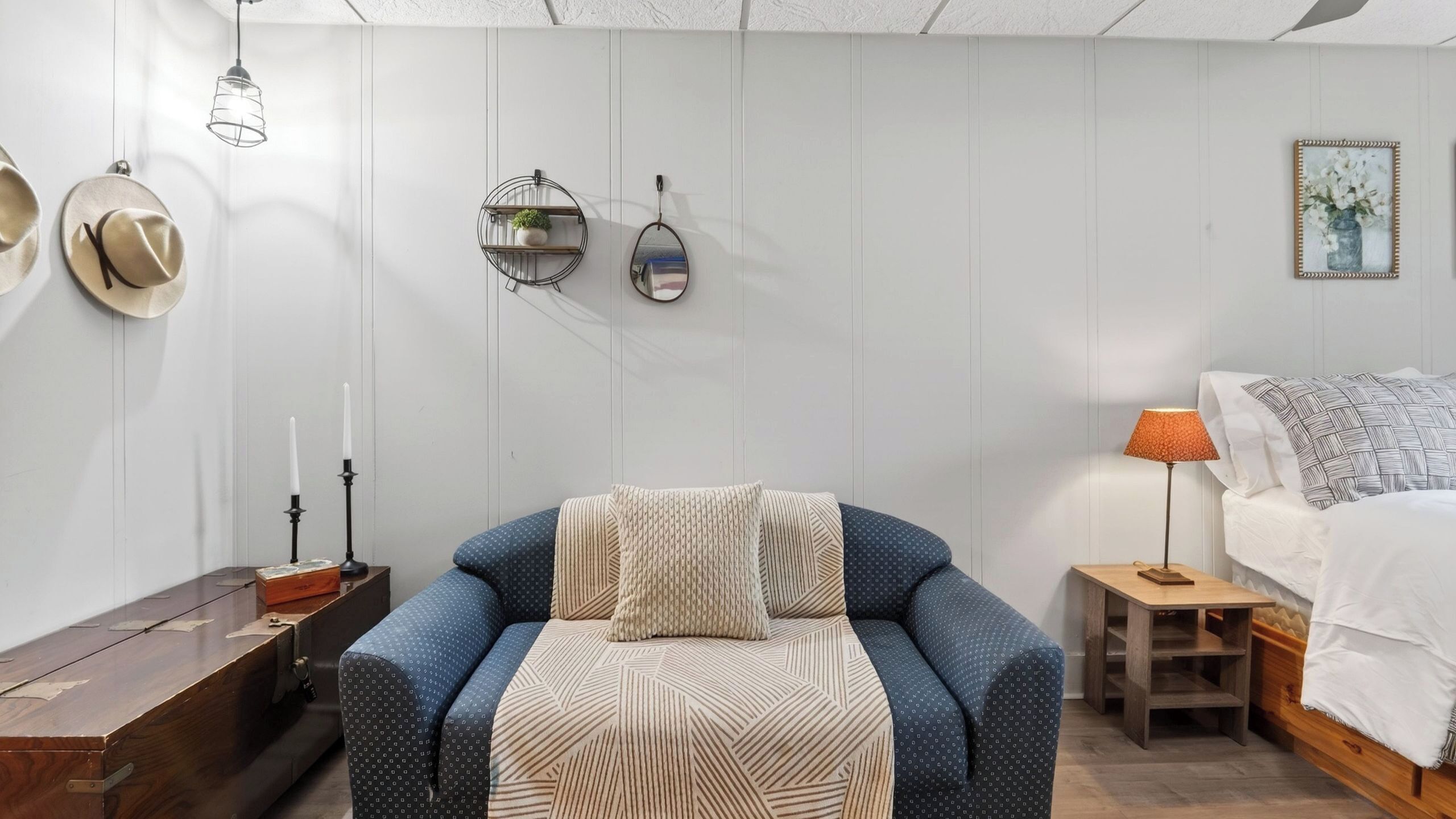 A bedroom with a bed, a chair, a small table, a lamp, and wall decorations including hats and a framed picture.