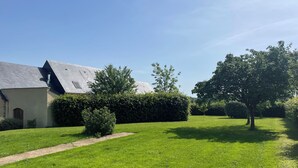 Een gazon met een gebouw, herkenbaar aan een schuin dak en schoorsteen, omgeven door bomen en heggen.