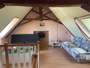Ein Schlafzimmer im Mansardenstil mit schrägem Dach, Holzbalken, einem Bett, einer Couch, einem kleinen Tisch und einem Regal mit Spielzeug.