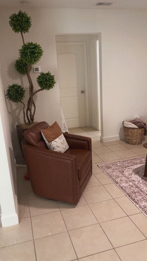 Een bruine leren fauteuil met een kussen met dessin, een kleine potplant en een vloerkleed met patroon op een betegelde vloer.