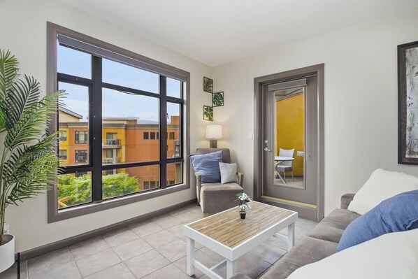 A modern living room with a large window, a sofa, a coffee table, and a door leading to a balcony. - Resort Living Across from the Lake: Pool, Hot Tub, Gym & 3BR !! (Kelowna)