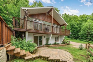 A two-story wooden cabin with a balcony, a stone pathway, and a forested backdrop.