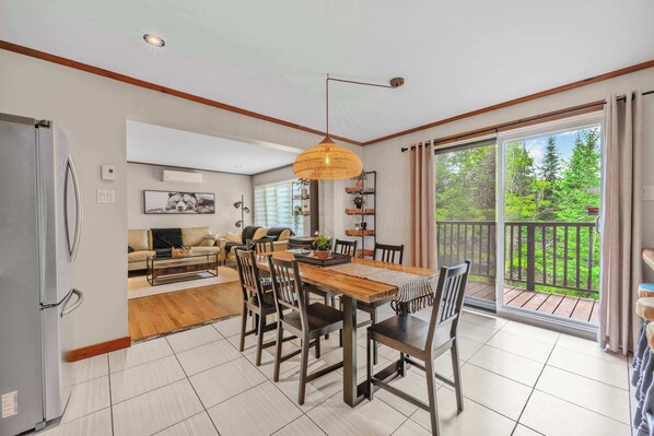 A spacious dining area with a wooden table and chairs, a refrigerator, and a large sliding glass door leading to a balcony with a view of greenery.