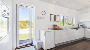 Una cocina moderna con un toldo de vidrio corredizo que da a un jardín, horno empotrado, un lavamanos con ventana y un reloj en la pared.
