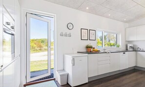 A modern kitchen with a sliding glass door leading to a garden, a built-in oven, a sink with a window, and a clock on the wall. - 5 Star Holiday Home in Glesborg (Glesborg)