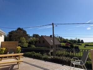 Een houten terras met een bankje en een stoel, een bakstenen huis op de achtergrond, en een metalen hek.
