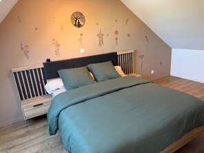 Een slaapkamer met een schuine kap, een bed met groen beddengoed, een houten nachtkastje, en een muur met bloemen- en boomstickers.