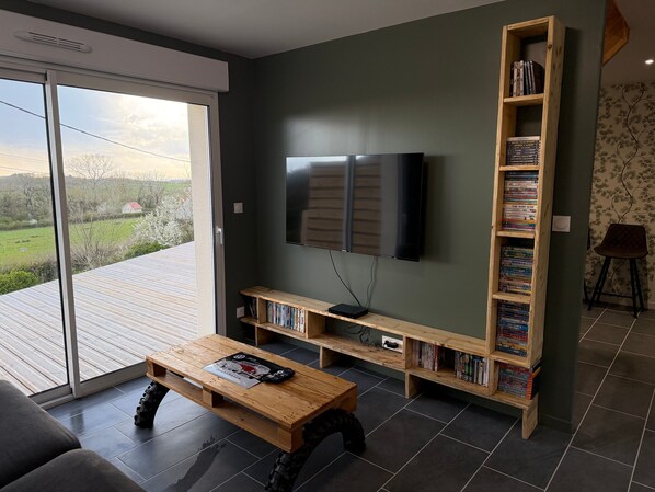 A living room with a TV, wooden shelves with books, a coffee table, and a view through a sliding door.