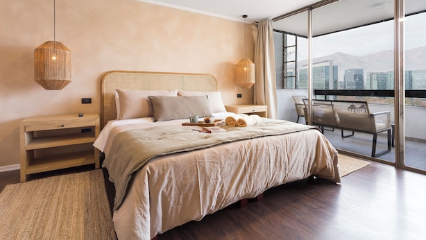 Een slaapkamer met een groot bed, houten hoofdbord, nachtkastjes, een nachttafeltje, een balkon met een tafel en stoelen, en uitzicht op de stad.