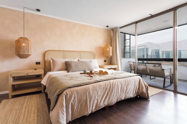 A bedroom with a large bed, wooden headboard, bedside tables, a nightstand, a balcony with a table and chairs, and a city view. - Spacious 4 Bedroom Apartment near Arauco Park (Santiago)