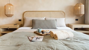 Een net opgemaakt bed met een tijdschrift, een schaal fruit en een afstandsbediening op een nachtkastje van hout.