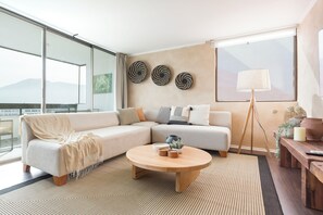 A modern living room with a sectional sofa, a round coffee table, and a large window with a view of mountains. - Spacious 4 Bedroom Apartment near Arauco Park (Santiago)