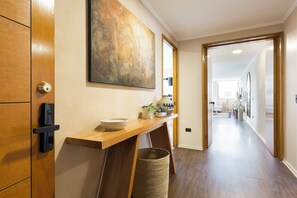 A hallway with a wooden door, a wall-mounted abstract painting, a wooden console table with a bowl, a plant, and a woven basket. - Spacious 4 Bedroom Apartment near Arauco Park (Santiago)