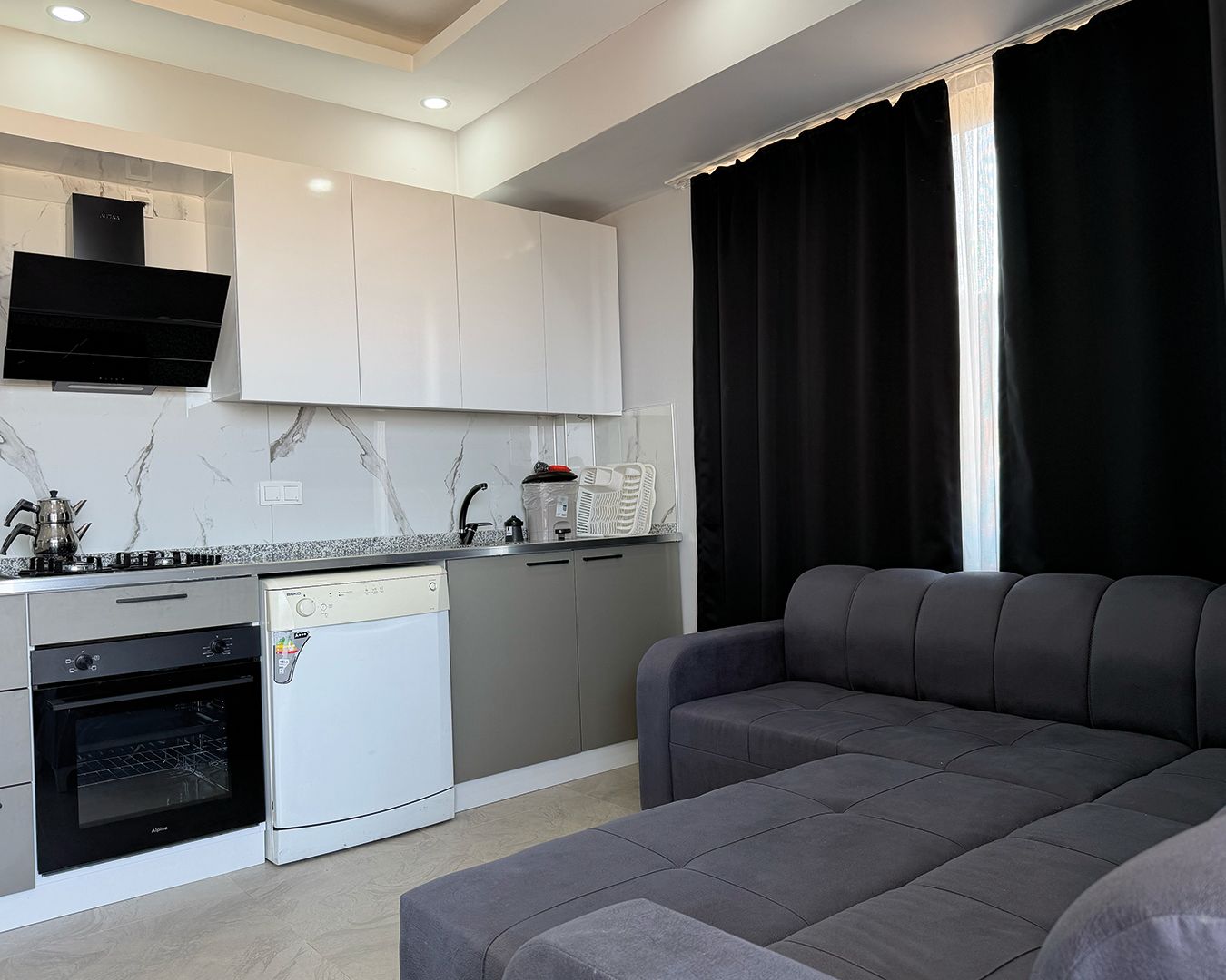 Una cocina moderna con horno empotrado, lavavajillas y estufa. Hay un sofá gris y una ventana con cortinas negras.
