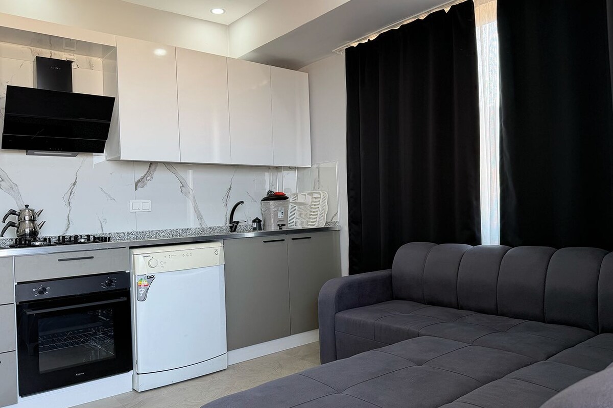 Una cocina moderna con horno empotrado, lavavajillas y estufa. Hay un sofá gris y una ventana con cortinas negras.