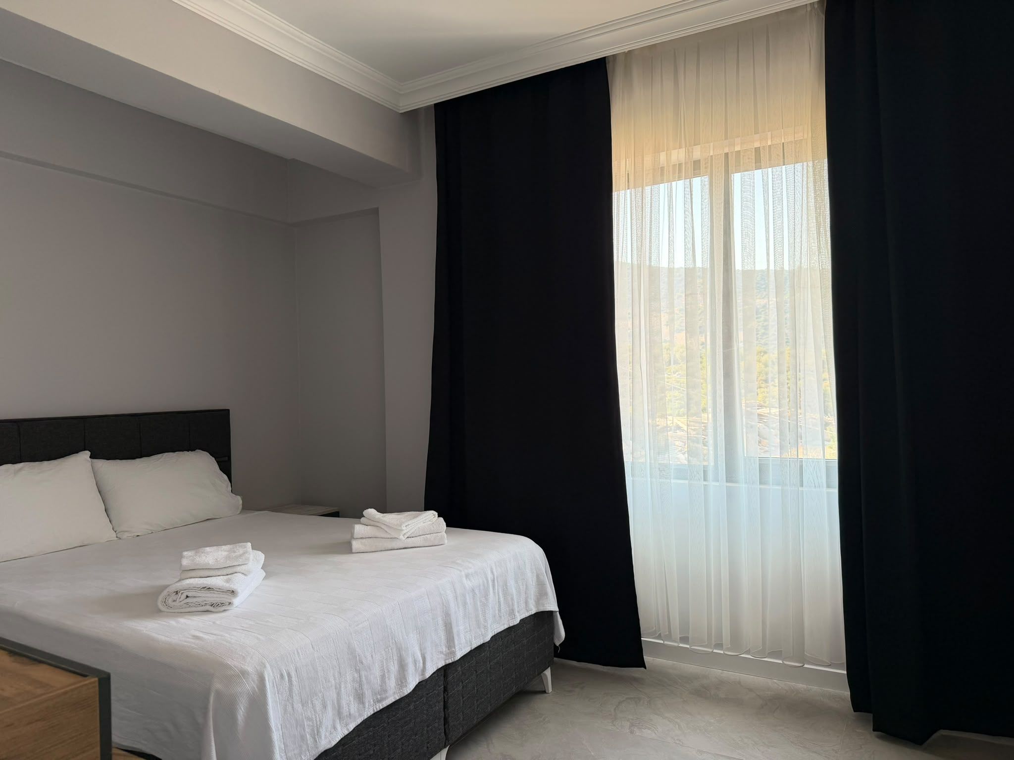 Une chambre d’hôtel avec un lit, des oreillers, une table de chevet et une fenêtre avec des rideaux légers.