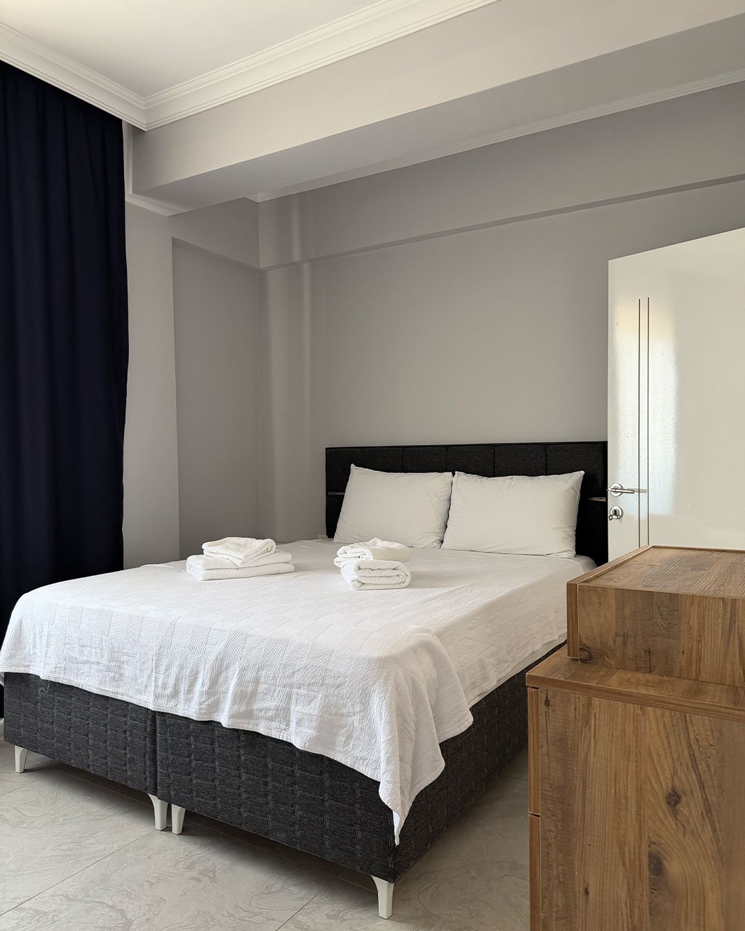 A neatly made bed with white linens and towels, a dark headboard, and a wooden nightstand to the right.