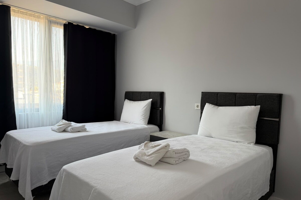 Dos camas individuales con sábanas y toallas blancas, una ventana con cortinas traslúcidas y una pared lisa.