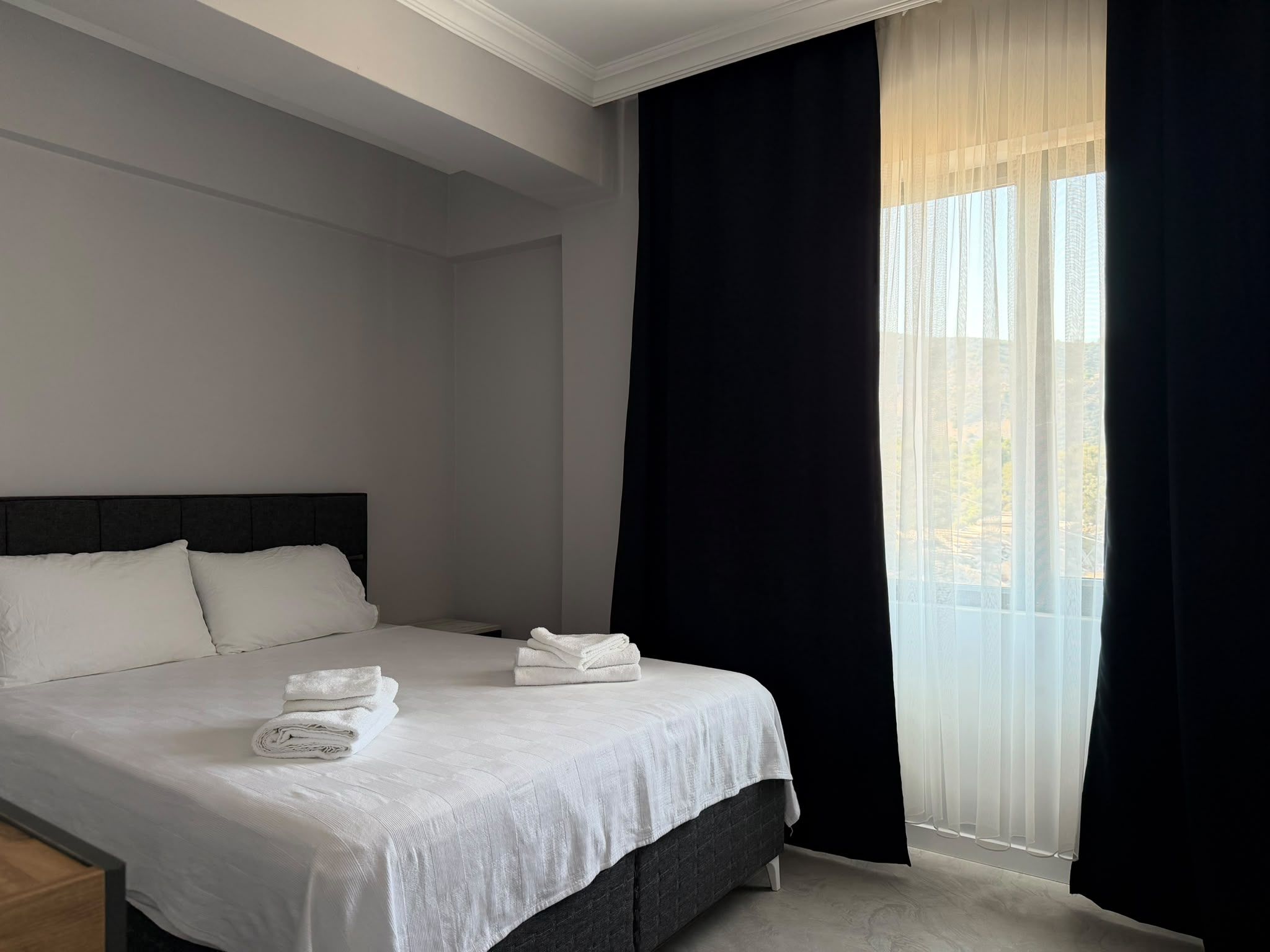 A hotel room with a bed, pillows, a nightstand, and a window with sheer curtains.