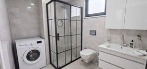 Un baño moderno con ducha envuelta en vidrio, lavadora, inodoro y lavamanos con artículos de aseo.