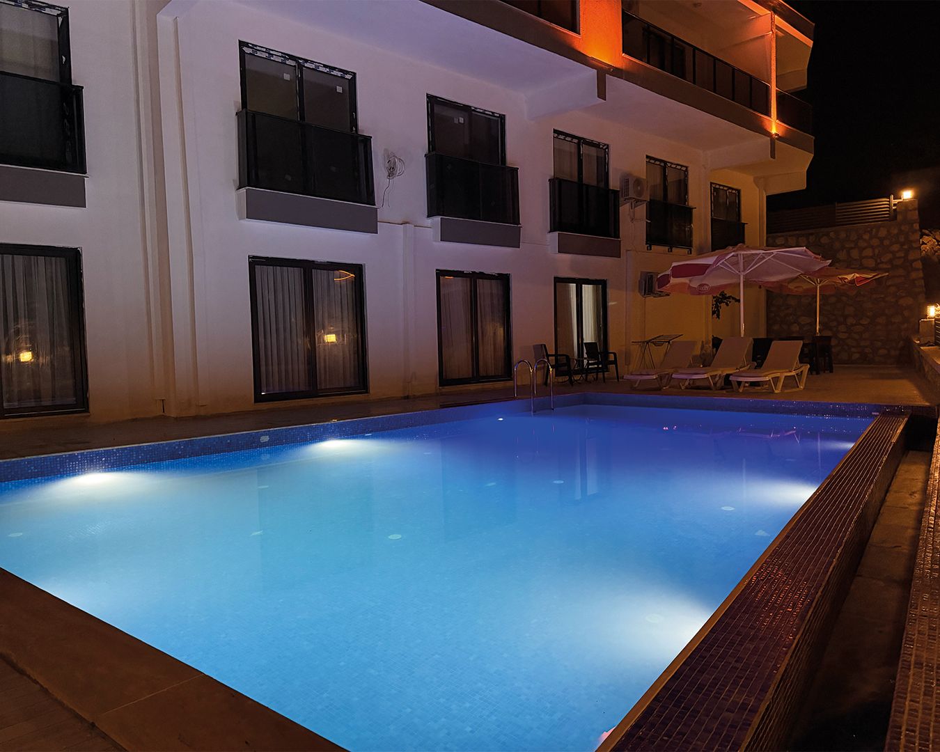 Una piscina con borde de azulejos, junto a un edificio con varias ventanas y una pared de piedra.