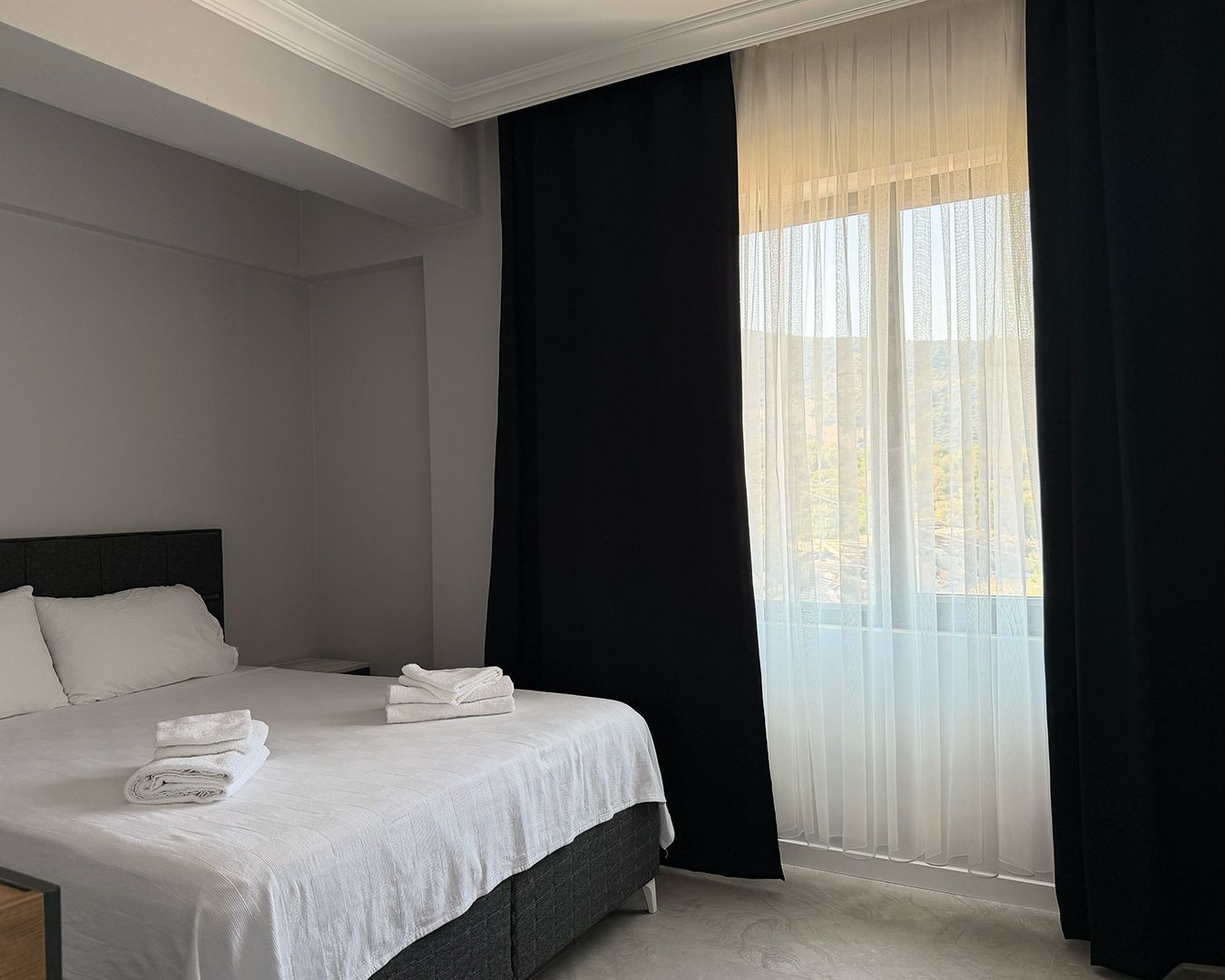 Una habitación de hotel con cama, mesita de noche y ventana con cortinas translúcidas.