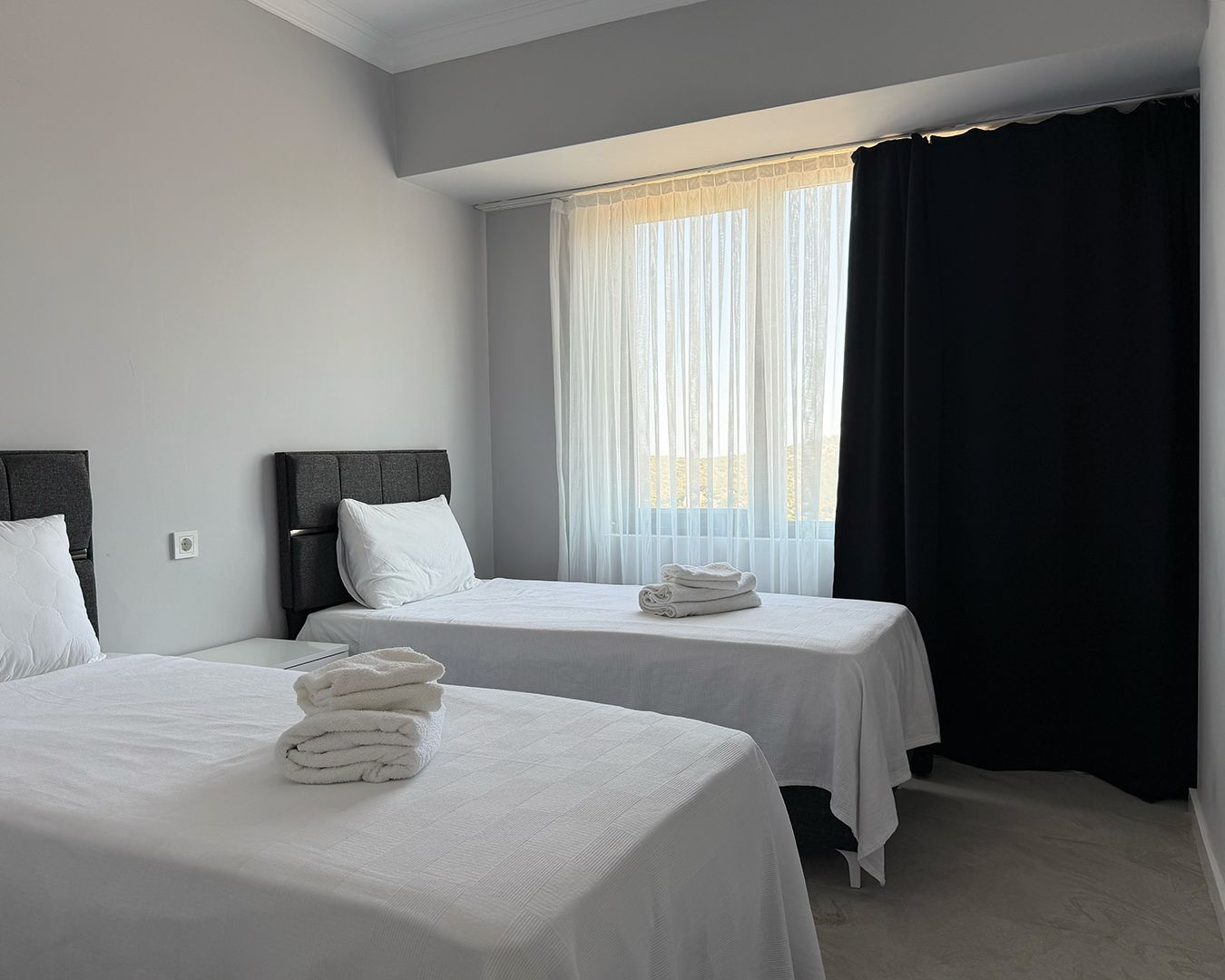 Habitación de hotel con dos camas, un ventanal con cortinas traslúcidas y una cortina oscura a la derecha.