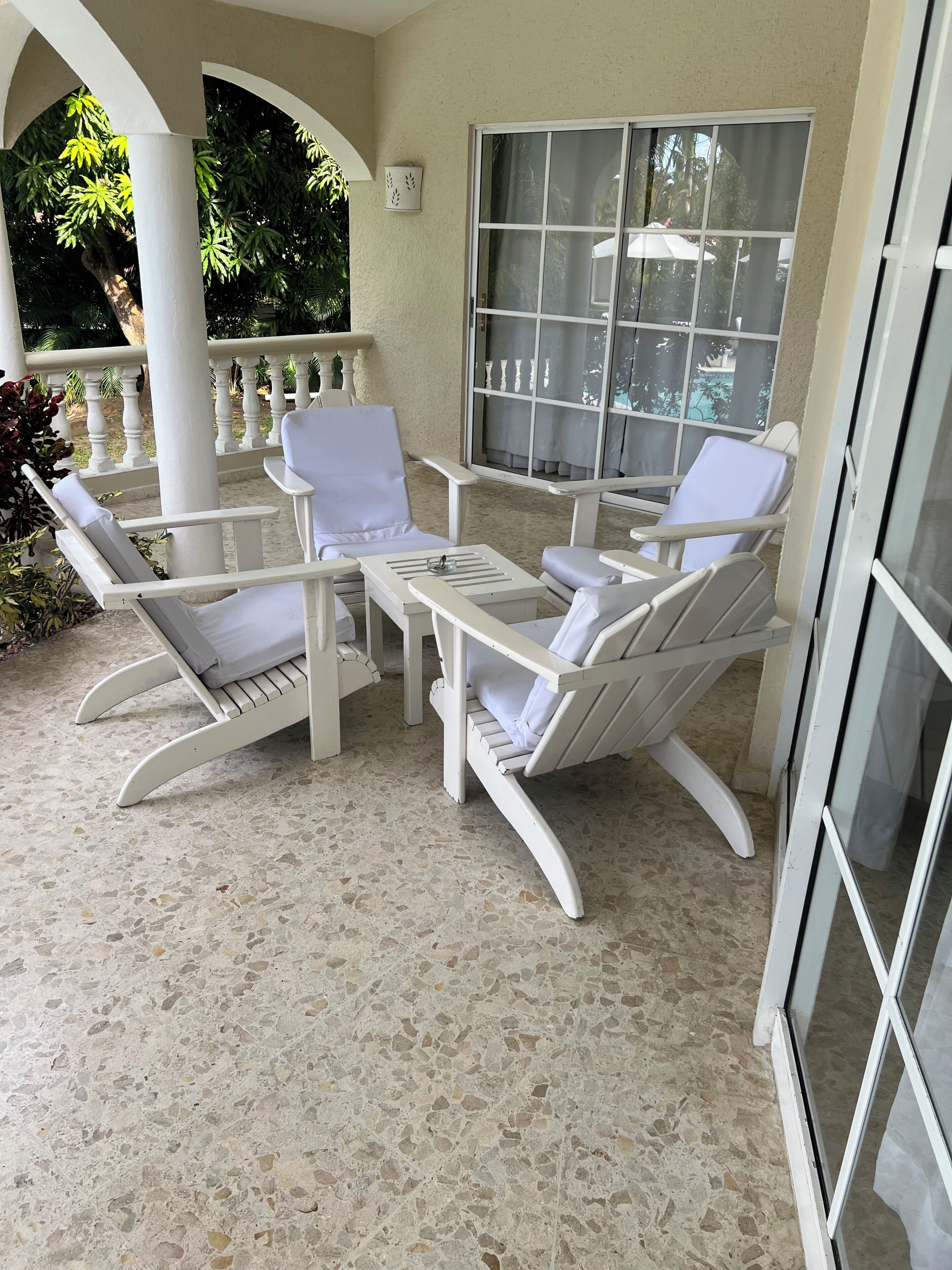 Een overdekt terras met witte lounge fauteuils en een tafel, een groot raam met gordijnen en een witte kolomconstructie.