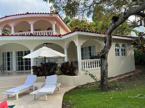 Ein zweistöckiges Gebäude mit einem Balkon, einem Poolbereich mit Liegestühlen und einem Baum im Vordergrund.