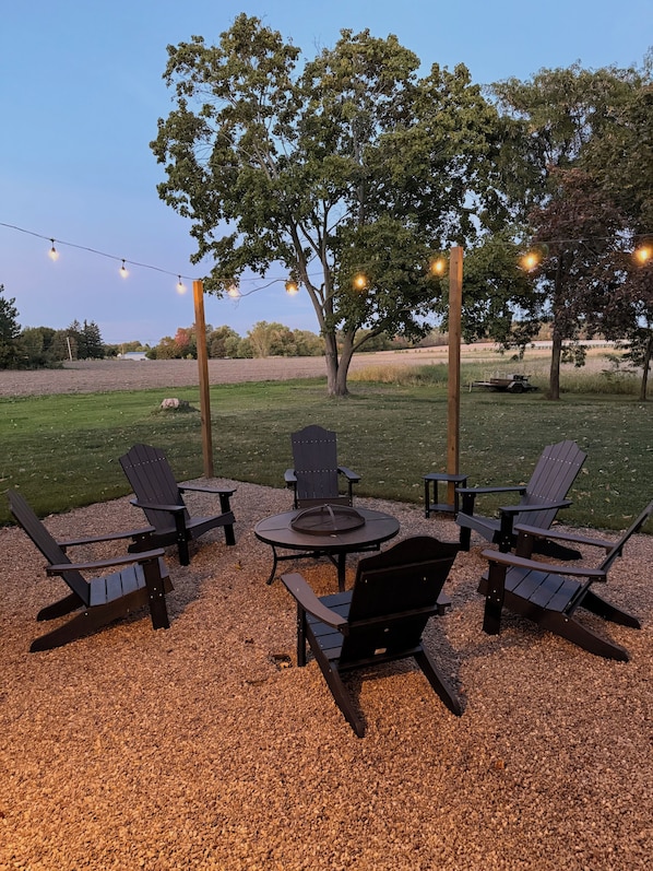 An outdoor seating area with Adirondack chairs and a round table, surrounded by gravel, under string lights.