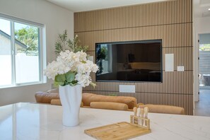 Et moderne køkken med en marmorbordplade, en træskærebræt, en hvid vase med blomster, et fladskærms-tv monteret på væggen og en beige sofa.