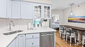 A kitchen with white cabinets, a stainless steel sink, and a dishwasher. There is a dining area with a wooden table and chairs. - Sundunes 173 (Navarre)