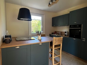 Cocina moderna con armarios oscuros, encimera de madera, lámpara colgante y ventana que deja entrar luz natural.