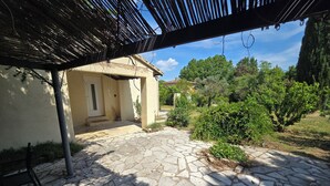 En stenbelagt terrasse med et pergola-dække, en dør og et lille haveareal med buske og træer.