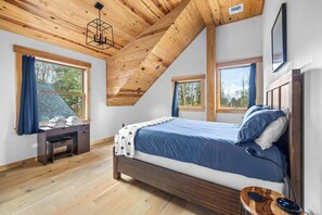 A bedroom with a wooden ceiling, a bed with blue bedding, a desk with a chair, and a window with a view of trees. - Spacious 4-BR Retreat Near Stratton & Mt. Snow (Stratton)