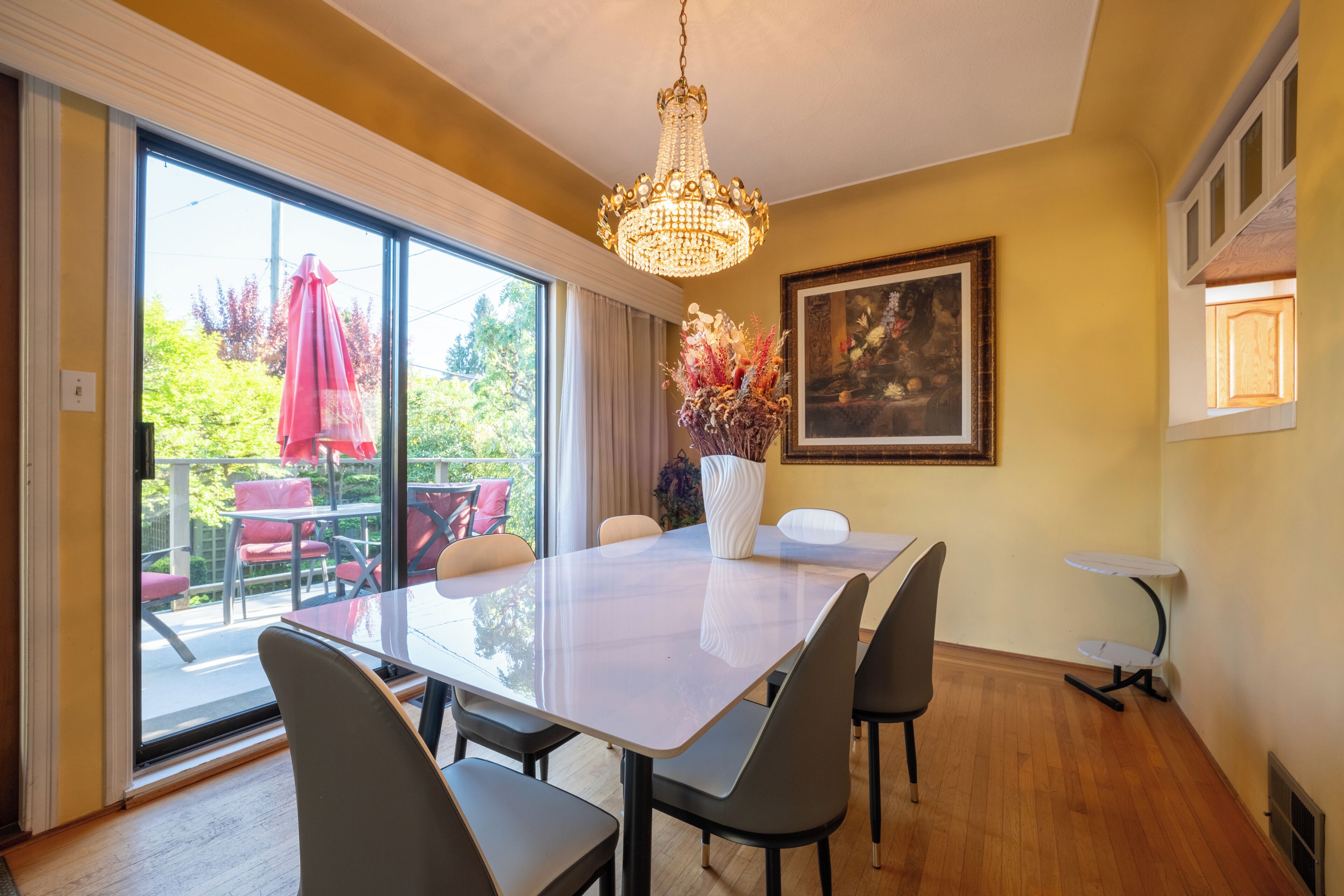A dining area with a large table, chairs, a chandelier, a painting, and a view through a sliding glass door.