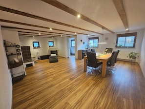 A spacious room with a wooden floor, a dining table with chairs, a black sofa, a TV on the wall, and a window with a view.