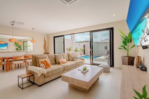 Ein modernes Wohnzimmer mit Sofa, Couchtisch, Essbereich und Blick auf einen Swimmingpool.