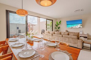 Ein modernes Wohnzimmer mit Essbereich, großen Fenstern, einer Couch und einem Fernseher, der eine Strandansicht zeigt.