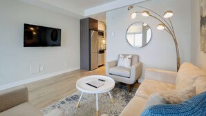 A modern living room with a flat-screen TV, a round mirror, a sofa, a coffee table, and a kitchen area visible through an open door. - Serene and Calming Downtown Suite (Victoria)