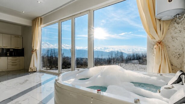 A modern bathroom with a large bathtub, a kitchen area, and a view of mountains through large windows.