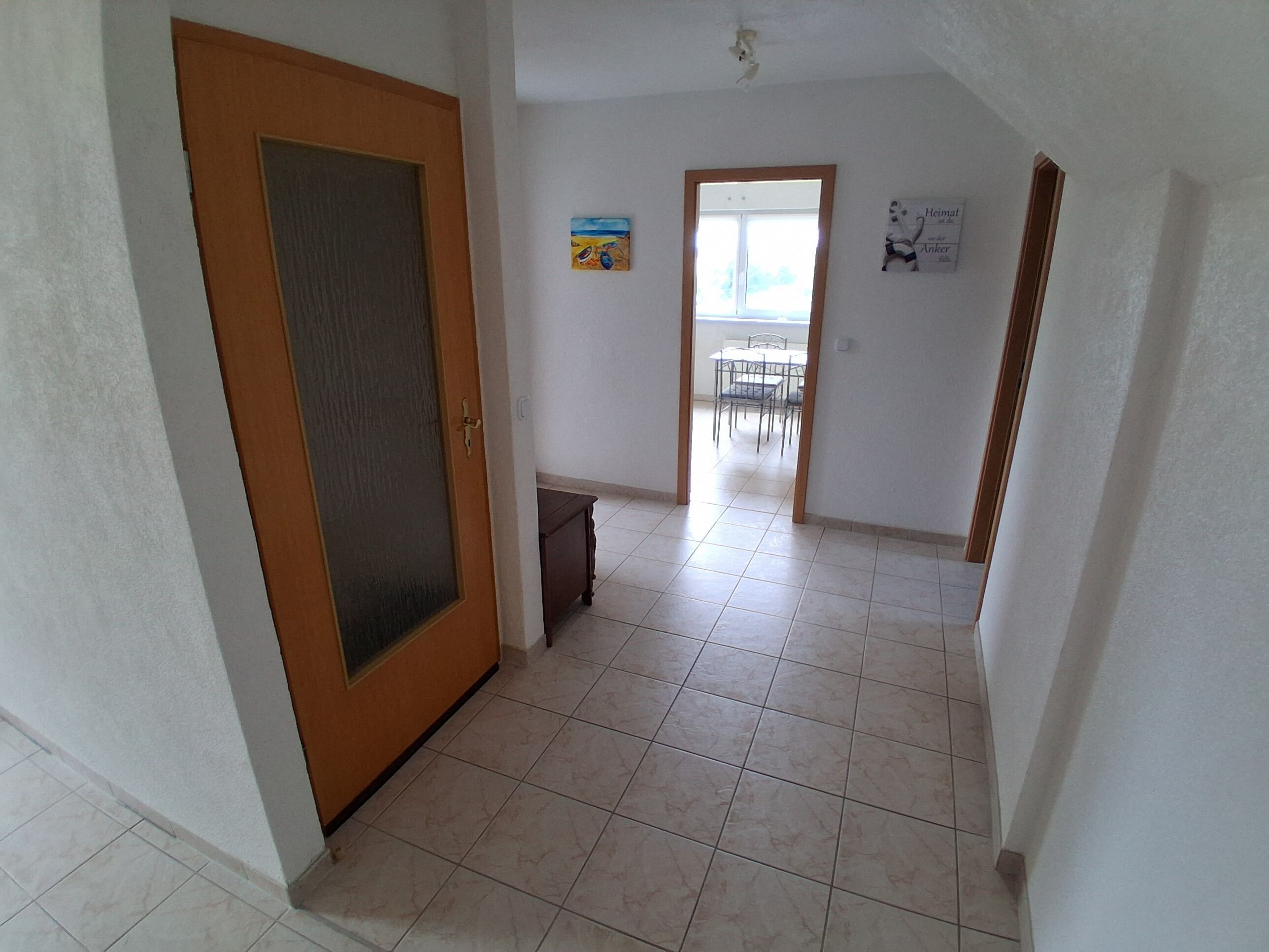Um corredor com porta de madeira, chão de azulejos, uma pequena lixeira e um espaço de jantar com cadeiras e uma mesa.