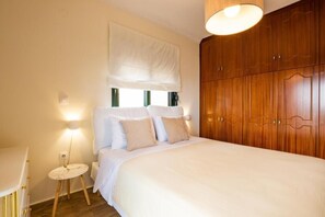 A bedroom with a bed, bedside table, lamp, wardrobe, and window with blinds. - Villa Maria (Karpathos)