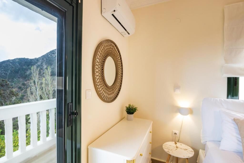 Una habitación con balcón, un aparador blanco, un espejo redondo, una mesita con lámpara y una cama.