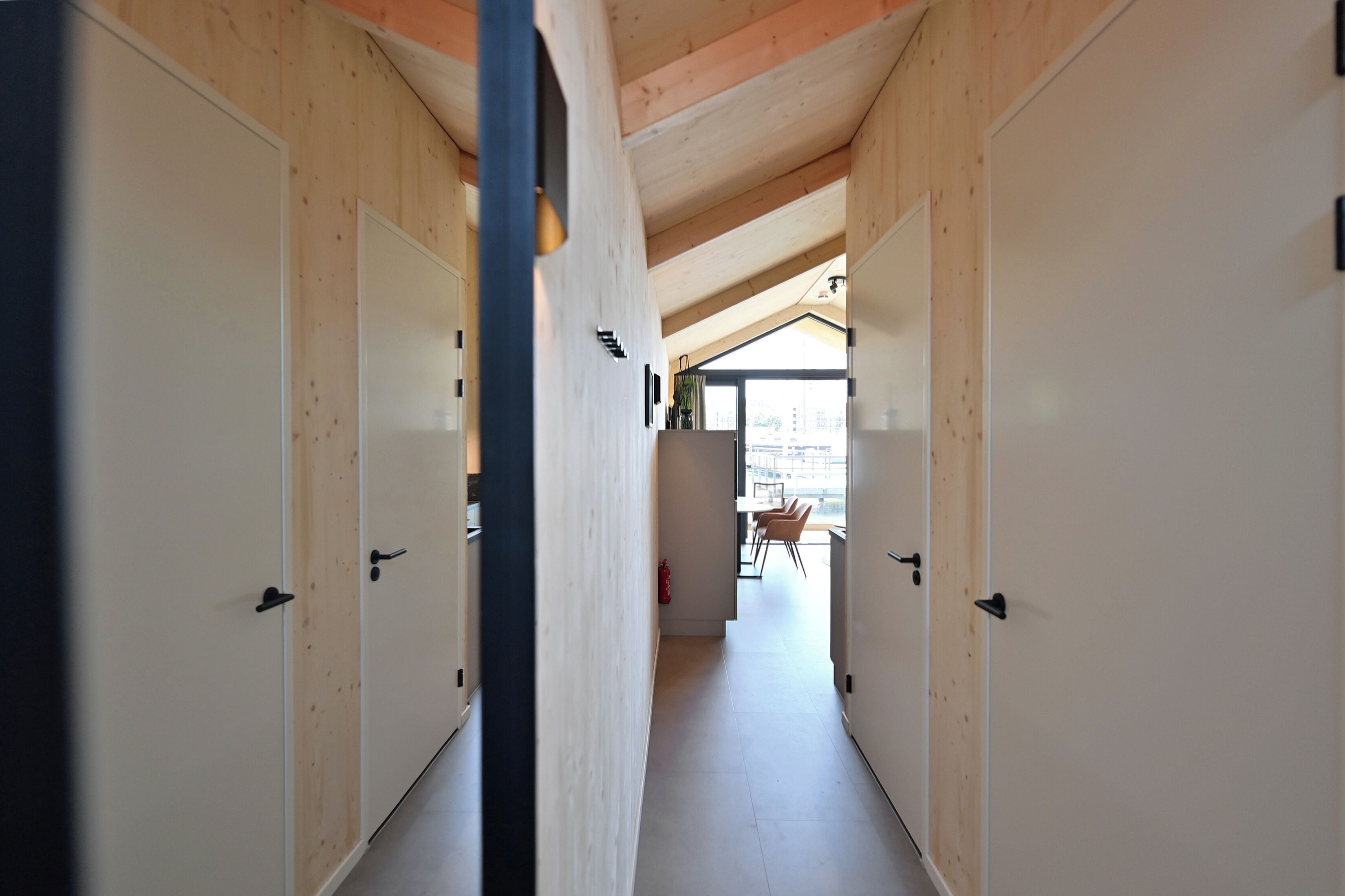 A corridor with wooden walls and doors, leading to a dining area with chairs and a table.