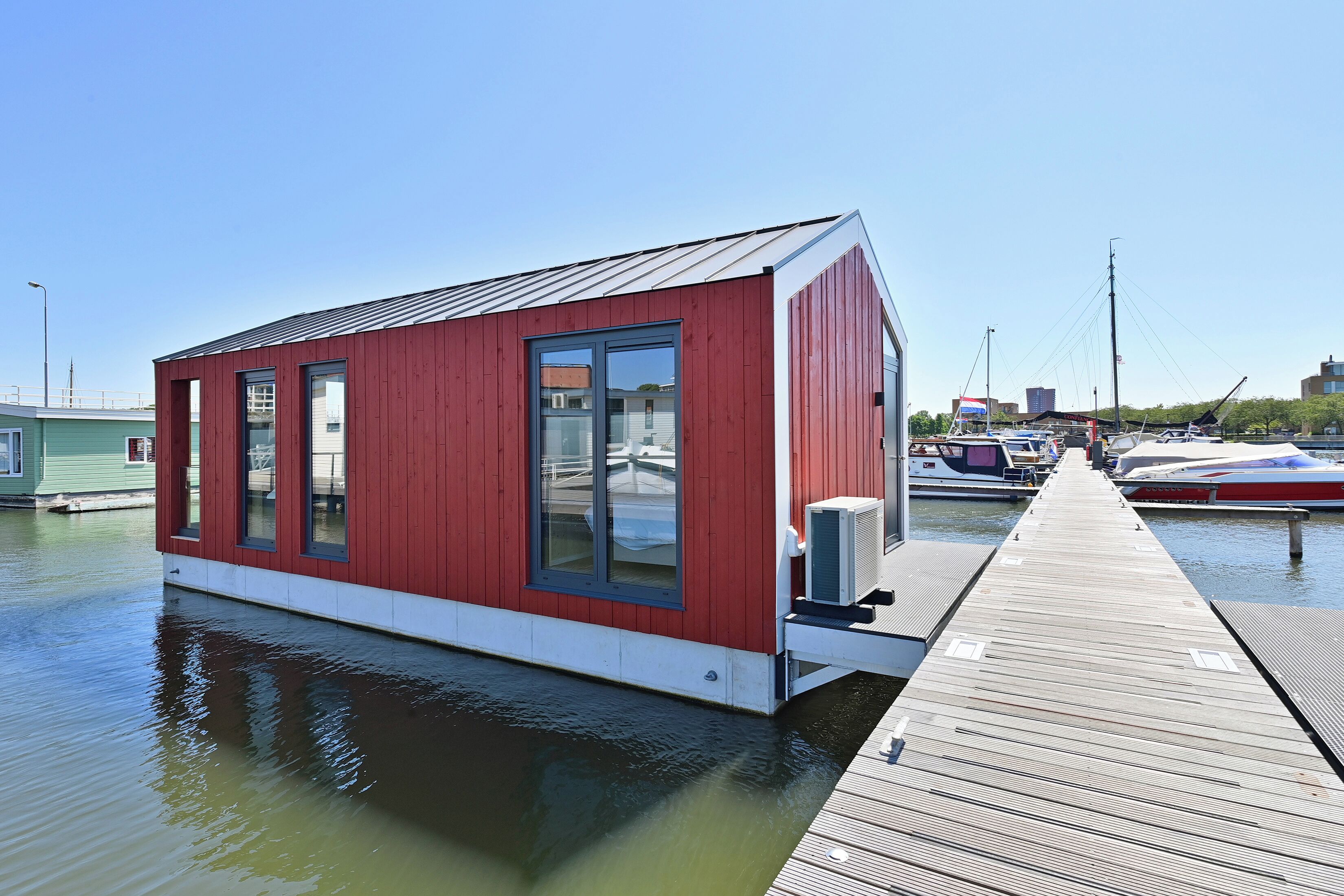 A red building with large windows, a dock extending into a body of water, and boats moored in the background.