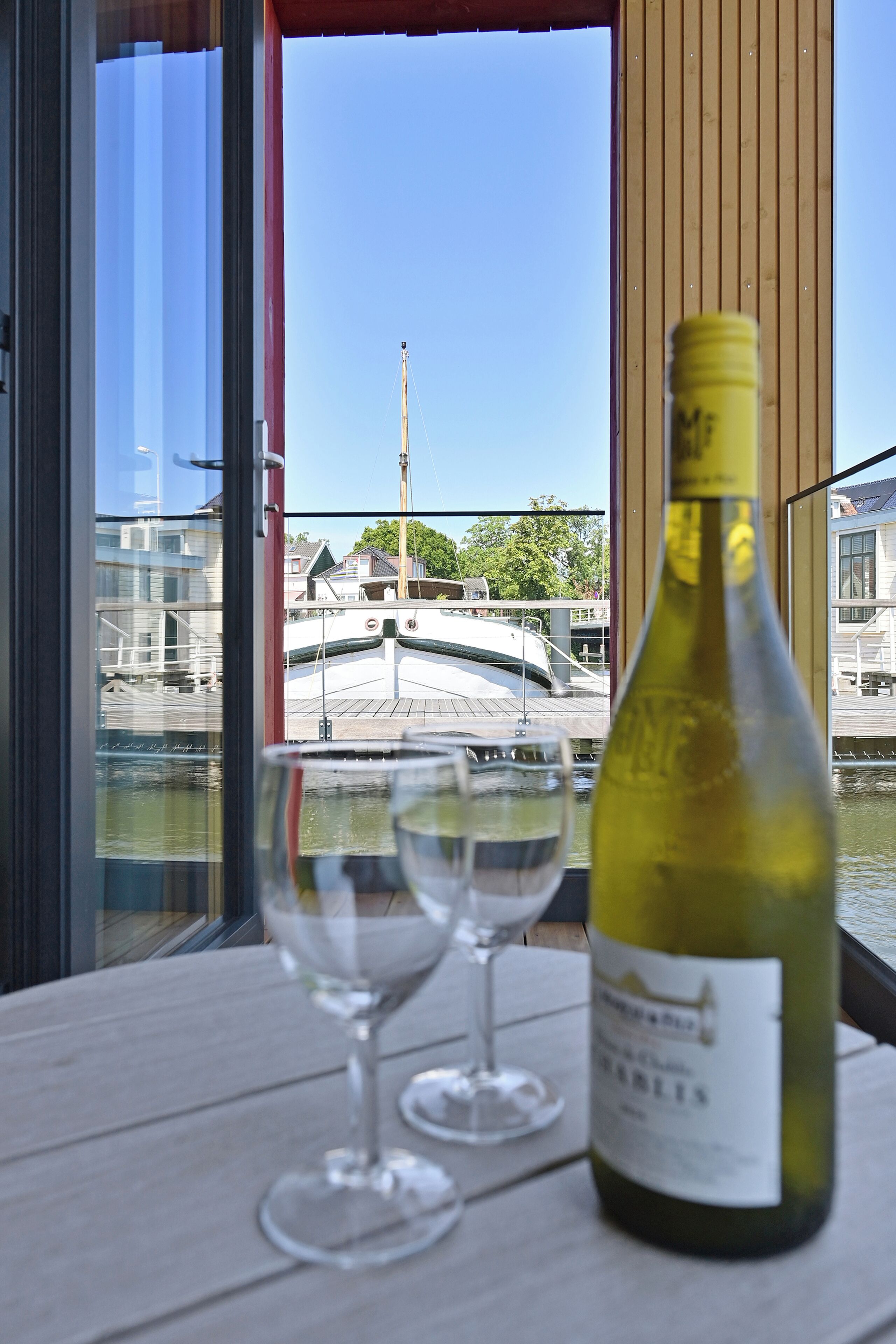 Una mesa con una botella de vino blanco y dos copas vacías, con vistas a un puerto deportivo con barcos y edificios.