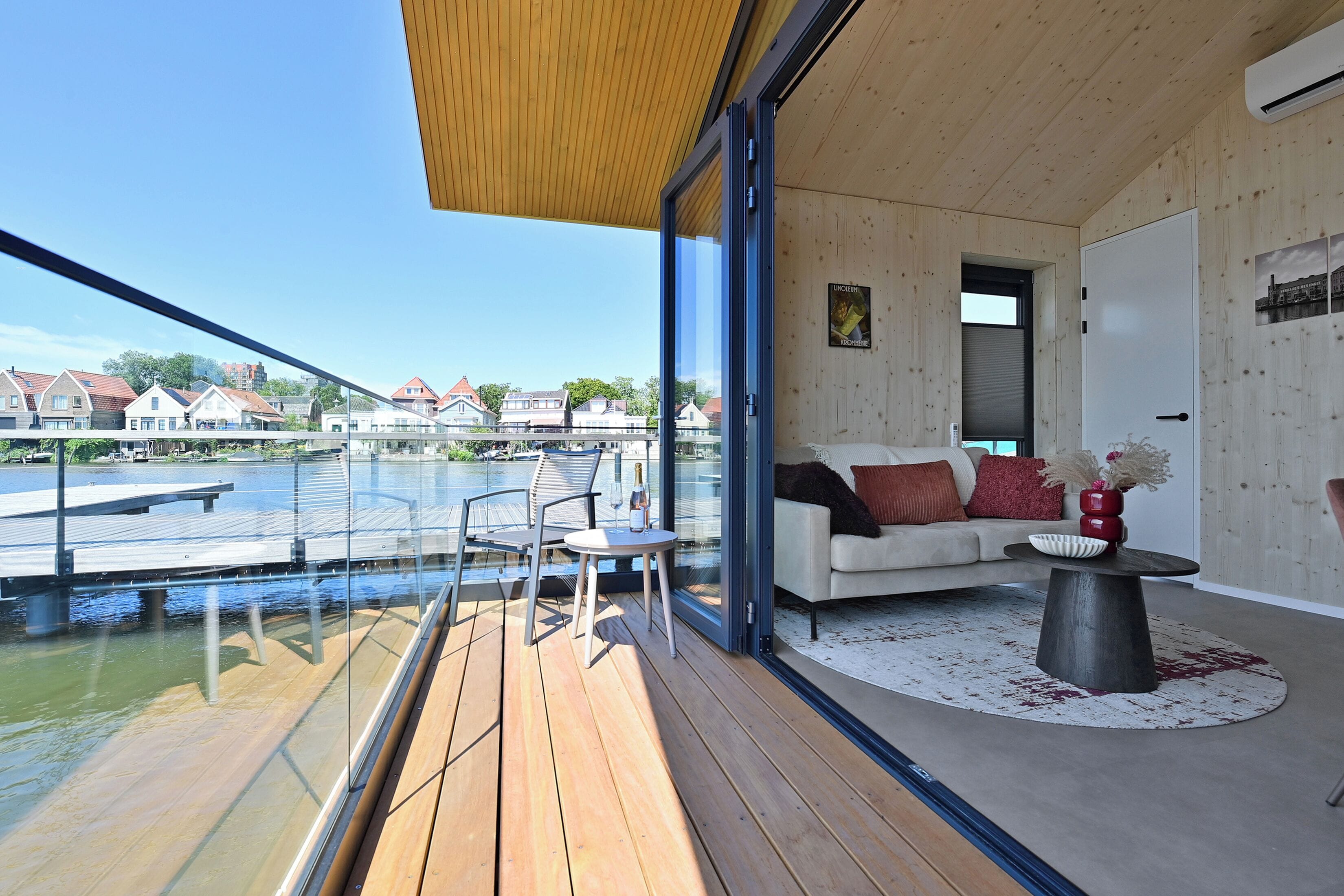 Sala moderna com guarda-corpo de vidro com vista para um corpo de água, um deck de madeira e casas ao longe.